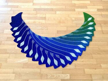 Gestrickter halbmondförmiger Schal mit blau-grünem Farbverlauf und weißen Federmotiven auf hellem Holzboden