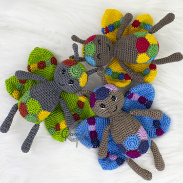 Drei gehäkelte Amigurumi‑Schmetterlinge mit bunten Kreisapplikationen auf weißem, flauschigem Hintergrund.