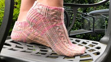 Füße in gestrickten kurzen Socken mit Zopfmuster in Rosa- und Grüntönen auf einem Metallstuhl