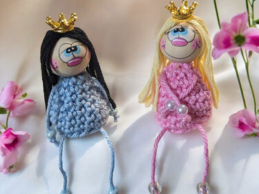 Zwei gehäkelte Prinzessinnenpuppen in Blau und Rosa mit Kronen und Perlen auf hellem Untergrund