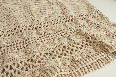 Close-up of beige crocheted fabric showing dense sections, lace rows, bobble stitches and a ribbed edge.