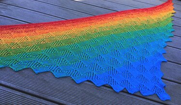 Gestricktes Dreieckstuch mit Regenbogen-Farbverlauf, feinem Lochmuster und gezacktem Rand auf dunklem Holz.