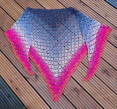 Gehäkeltes Dreieckstuch mit Farbverlauf (Blau–Weiß–Pink/Orange) auf Holzdielen