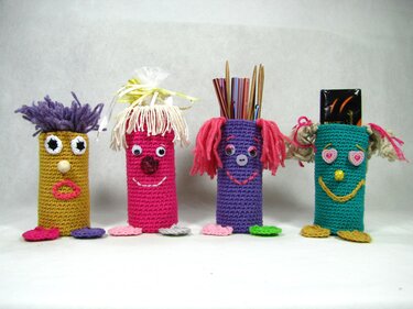 Vier gehäkelte Utensilos in Gelb, Pink, Lila und Türkis mit Gesichtern und kleinen Füßen