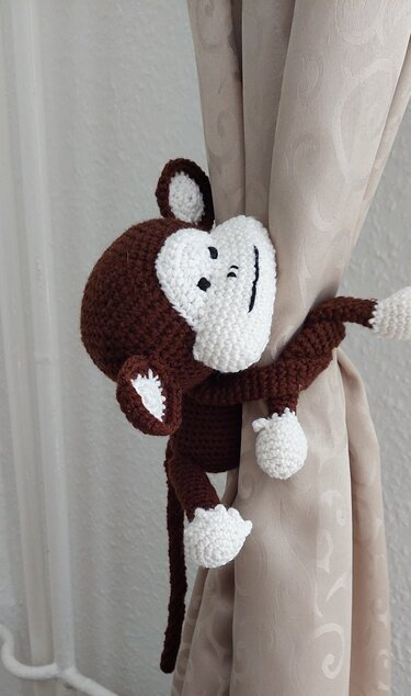 Gehäkelter braun-weißer Amigurumi-Affe umschlingt eine helle Gardine als Raffhalter.
