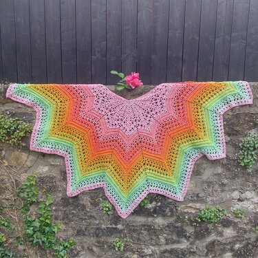 Star-shaped crochet shawl with pink-to-green gradient, draped on a stone wall with a pink rose