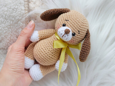 Close-up of a small brown crocheted amigurumi puppy with white paws, dark brown ears, and a yellow ribbon held in a hand