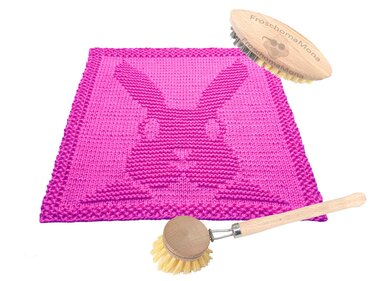 Quadratisches pinkes gestricktes Spültuch mit erhabenem Hasenkopf-Motiv; zwei Holzbürsten liegen daneben