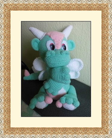 Gehäkelter Amigurumi-Drache in Mintgrün mit rosa und weißen Akzenten, sitzend.