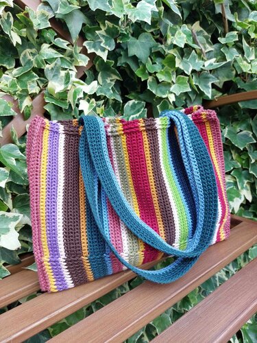 Gehäkelte Tasche mit bunten Streifen und blauen Henkeln auf Holzbank vor Efeu.