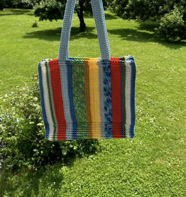 Gehäkelte Henkeltasche mit bunten vertikalen Streifen und hellblauen Henkeln vor grünem Rasen