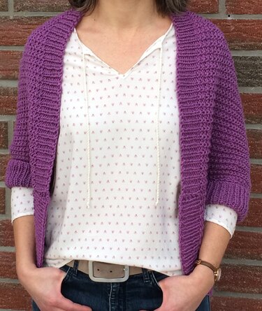 Frau trägt offenen lila Seelenwärmer mit 3/4-Ärmeln über weißer gemusterter Bluse und Jeans vor Backsteinwand