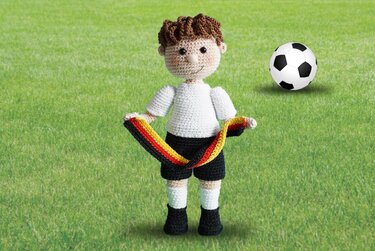 Gehäkelter Amigurumi-Junge in weißem Trikot mit schwarz‑rot‑goldenem Schal neben einem Fußball auf Rasen.