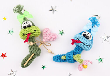 Two crocheted snake keychains on a white background with star confetti — green with a pink heart and blue with a small charm.