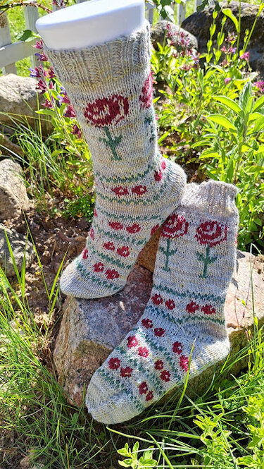Gestrickte graubeige Socken mit roten Blumenmotiven und grünen Stängeln; eine Socke auf einem Formfuß, die andere daneben auf einem Stein