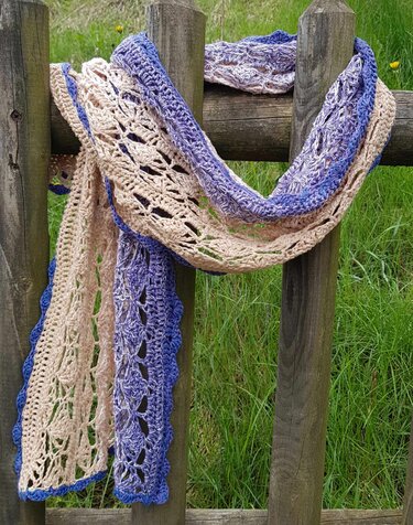 Gehäkeltes Rechtecktuch in Beige und Violett mit blauem Rand, drapiert über einen Holzzaun vor Gras