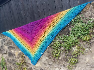 Triangle crochet shawl in rainbow gradient with lace rows, draped on a stone wall against a dark wooden fence