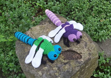 Zwei farbenfrohe Amigurumi‑Libellen (grün‑blau und lila‑rosa) auf einem Stein.