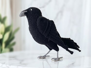 Black crocheted crow amigurumi with grey beak and grey legs standing on a marble surface