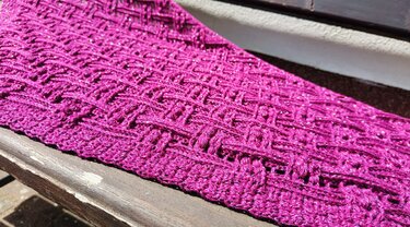Close-up of fuchsia crochet triangle shawl with raised openwork stitch, ribbed border, and sparkly yarn on a wooden bench