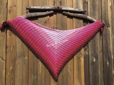 Gehäkeltes Dreieckstuch mit Pink-Farbverlauf und Lochreihenmuster an einer Holzwand aufgehängt.