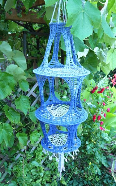 Gehäkeltes dreistufiges Hängeutensilo in Blau mit weißen Einsätzen, an Rankgerüst hängend.