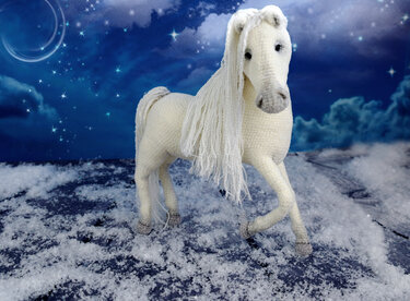 White crocheted amigurumi horse with long yarn mane and tail standing on a snowy surface under a deep blue starry sky with a crescent moon.