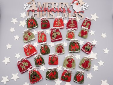 Nahaufnahme vieler kleiner rot-grüner gehäkelter Säckchen mit Weihnachtsmotiven, arrangiert um ein hölzernes Weihnachtsschild und weiße Sterne.