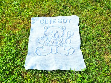 Hellblaue gehäkelte Babydecke mit reliefartigem Teddybär und Schriftzug 'CUTE BOY' auf Gras