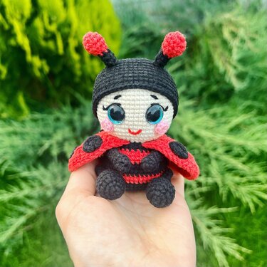 Hand holding a kawaii-style crocheted ladybug amigurumi with red wings, black body, glossy eyes and pink cheeks against green foliage