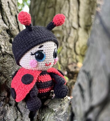 Red and black crocheted amigurumi ladybug doll with large glossy eyes sitting on tree bark