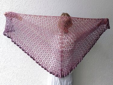 Person hält ein großes, gehäkeltes Dreieckstuch mit offenem Netz- und Farbverlauf in Lila- und Violetttönen hoch.
