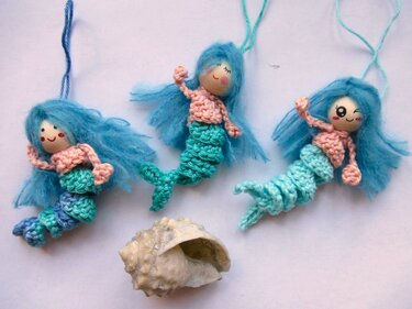 Drei gehäkelte Amigurumi-Meerjungfrauen mit blauen Haaren und pastellfarbenen Schwänzen neben einer Muschel auf hellem Hintergrund.