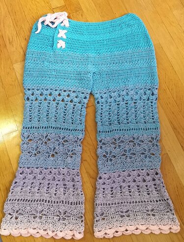 Gehäkelte Damenhose in Türkis bis Lila mit offenem Muster und Muschelsaum, liegend auf Holzboden