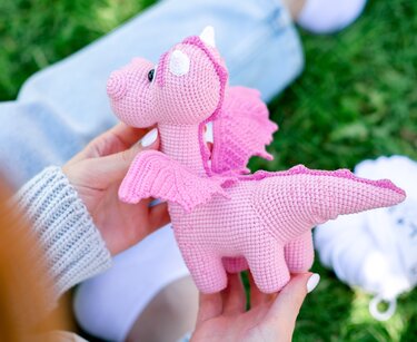 Small pink crochet amigurumi dragon with scalloped wings and white horns held in hands on grass