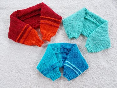 Drei Paare gestrickter Puppen-Armstulpen in Rot-Orange, Türkis und Blau auf hellem Untergrund.