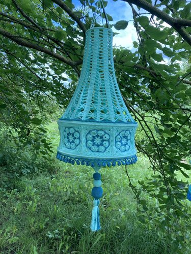 Türkis-blaue gehäkelte Hängedekoration in Glockenform mit Lochmuster, Grannysquares und Quaste, an einem Ast hängend.