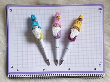 Drei kleine, handgehäkelte Wichtel mit weißen Bärten und Marienkäfern als Stiftüberzüge auf einem karierten Spiralblock