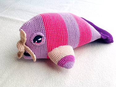 Gehäkelter Amigurumi-Wal in gestreiften Lila- und Pinktönen mit kleinen Flossen auf hellem Untergrund.