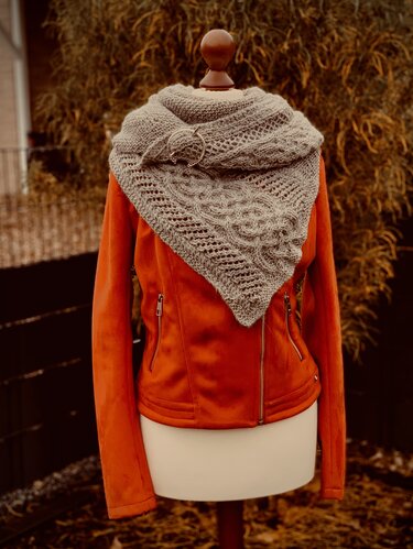 Gray knitted triangle shawl with cable and lace panels pinned with circular shawl pin on mannequin wearing orange jacket