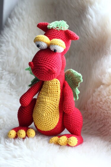 Red crocheted dragon amigurumi with yellow belly and green wings on a white fluffy background.