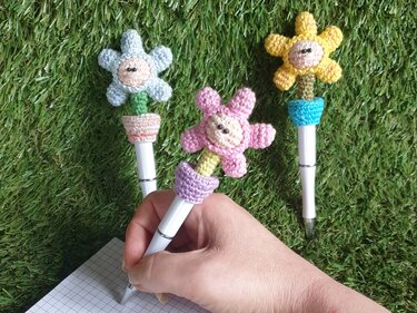 Nahaufnahme von drei weißen Stiften mit gehäkelten Blumenaufsätzen in Pastellfarben; eine Hand hält den mittleren Stift über kariertem Papier
