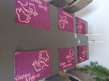 Gehäkelte Tischsets in brombeerfarben mit Kochmützen- und ",Guten Appetit"-Motiven auf einem Esstisch.