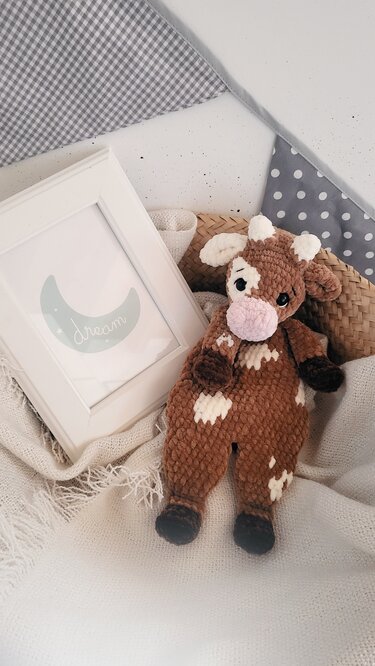 Braun-weißes gehäkeltes Kuh-Kuscheltier mit rosa Schnauze auf einer hellen Decke neben einem Bilderrahmen mit Mondmotiv.