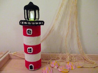 Handgehäkelter rot-weißer Leuchtturm mit schwarzer Laterne und LED-Kerze, neben Fischernetz und gehäkelten Muscheln.