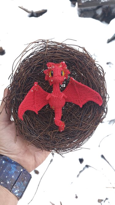 Red crocheted dragon amigurumi with yellow eyes and bead accents in a twig nest held over snow.