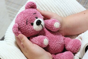 Pink crocheted amigurumi teddy bear with white snout and paw pads held in hands on a cream knit blanket