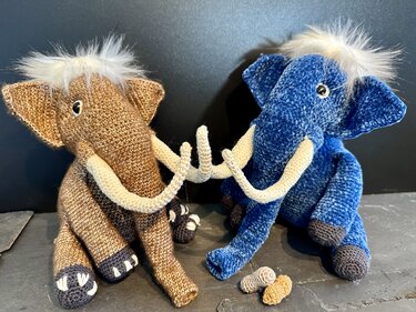 Zwei gehäkelte Amigurumi-Elefanten, einer braun, einer blau, mit hellen Stoßzähnen und weißer Mähne