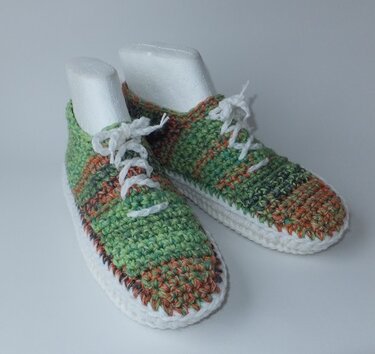 Gehäkelte Sneaker-Hausschuhe, grün-braun meliert, mit weißen Schnürsenkeln und weißer Sohle auf hellem Hintergrund.