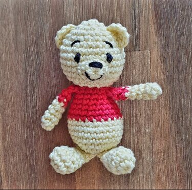 Mini-Amigurumi-Teddybär aus cremefarbenem Garn mit rotem Oberteil auf Holzuntergrund